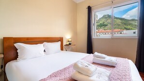 Ein Hotelzimmer mit einem Bett, einem Nachttisch, einer Lampe, einem Fenster mit Blick auf eine hügelige Landschaft und Handtüchern auf dem Bett.