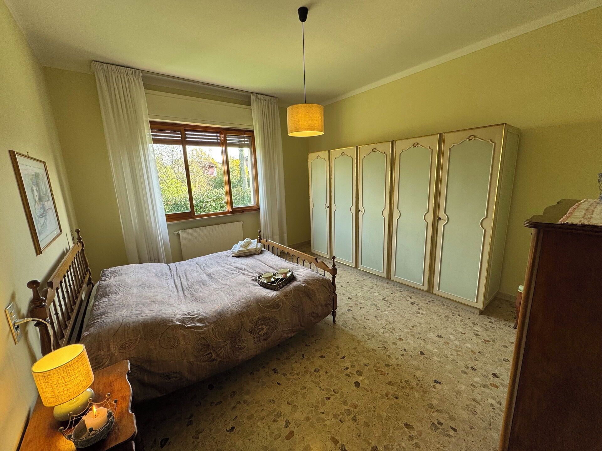 A bedroom with a bed, a bedside table with a lamp, a window with curtains, a radiator, and a wardrobe.