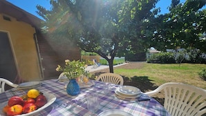 Un tavolo da pranzo all'aperto con tovaglia a quadri, sedie bianche, una ciotola di frutta, un vaso con fiori e un bicchiere d'acqua.