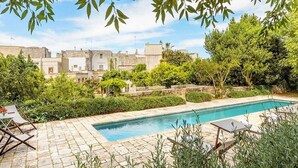 En swimmingpool med liggestole og en stenbelagt terrasse, omgivet af grøn vegetation og bygninger.