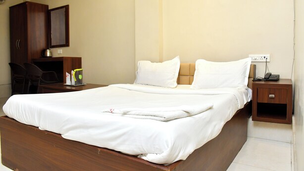 A neatly made bed with white linens, a wooden nightstand with a phone, and a small desk with a chair and a kettle. - Raj Gharana (Raipur)