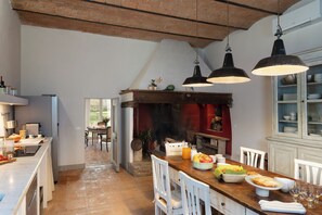 Una cucina spaziosa con soffitto in mattoni rustici, un'isola in legno con frutta e pane, e un'area pranzo con sedie bianche.