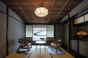Une pièce de style japonais traditionnel, avec un sol recouvert de tatamis, une table basse, deux chaises, une lanterne suspendue et une vue sur un bâtiment traditionnel à travers un paravent en papier.