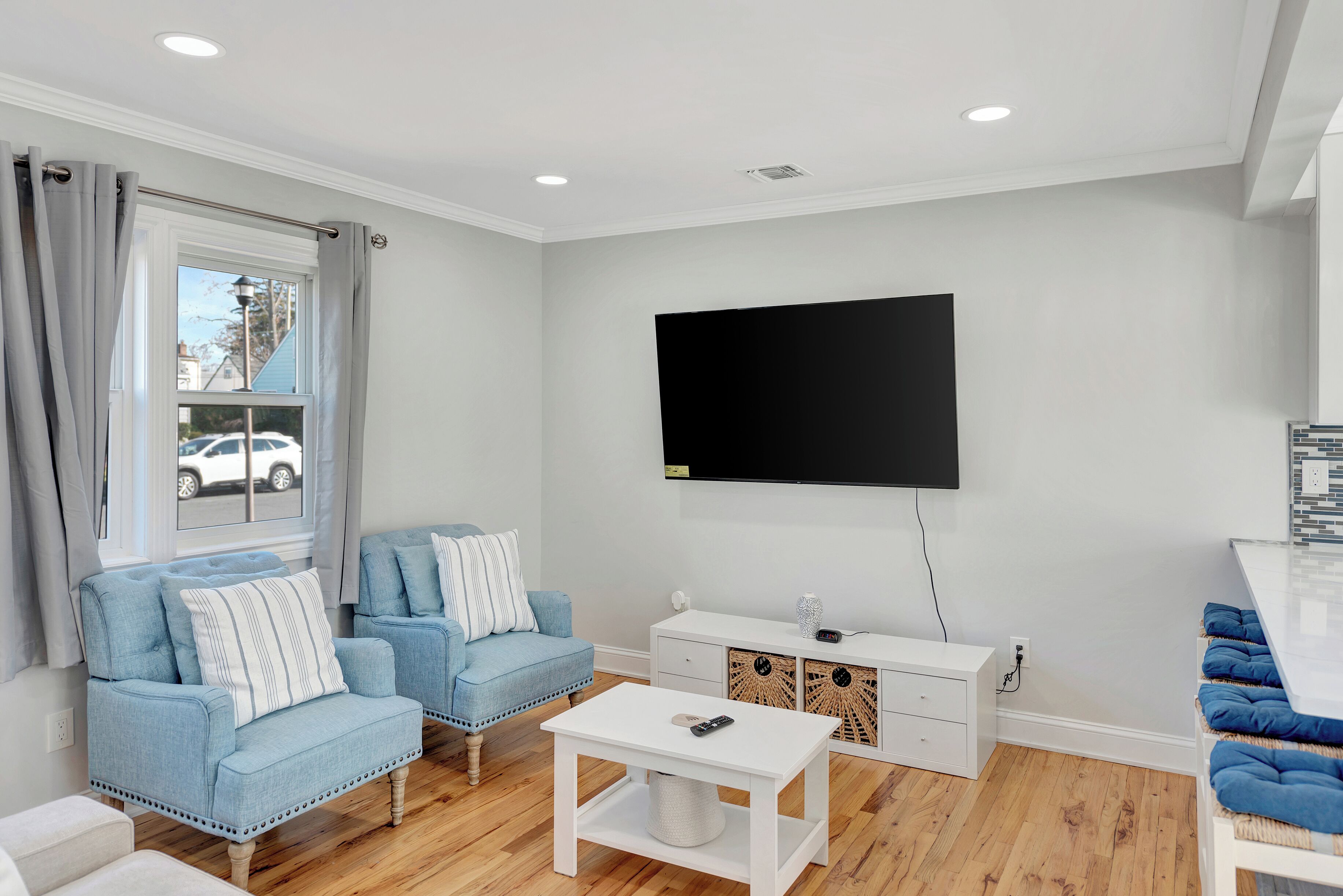 A modern living room with a flat-screen TV mounted on the wall, a white coffee table, two blue sofas with striped pillows, and a white cabinet with wicker baskets.