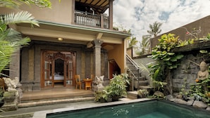 A pool area with a stone wall, a wooden deck, and a staircase leading to a balcony. - Togog Polos Ubud House (Ubud)