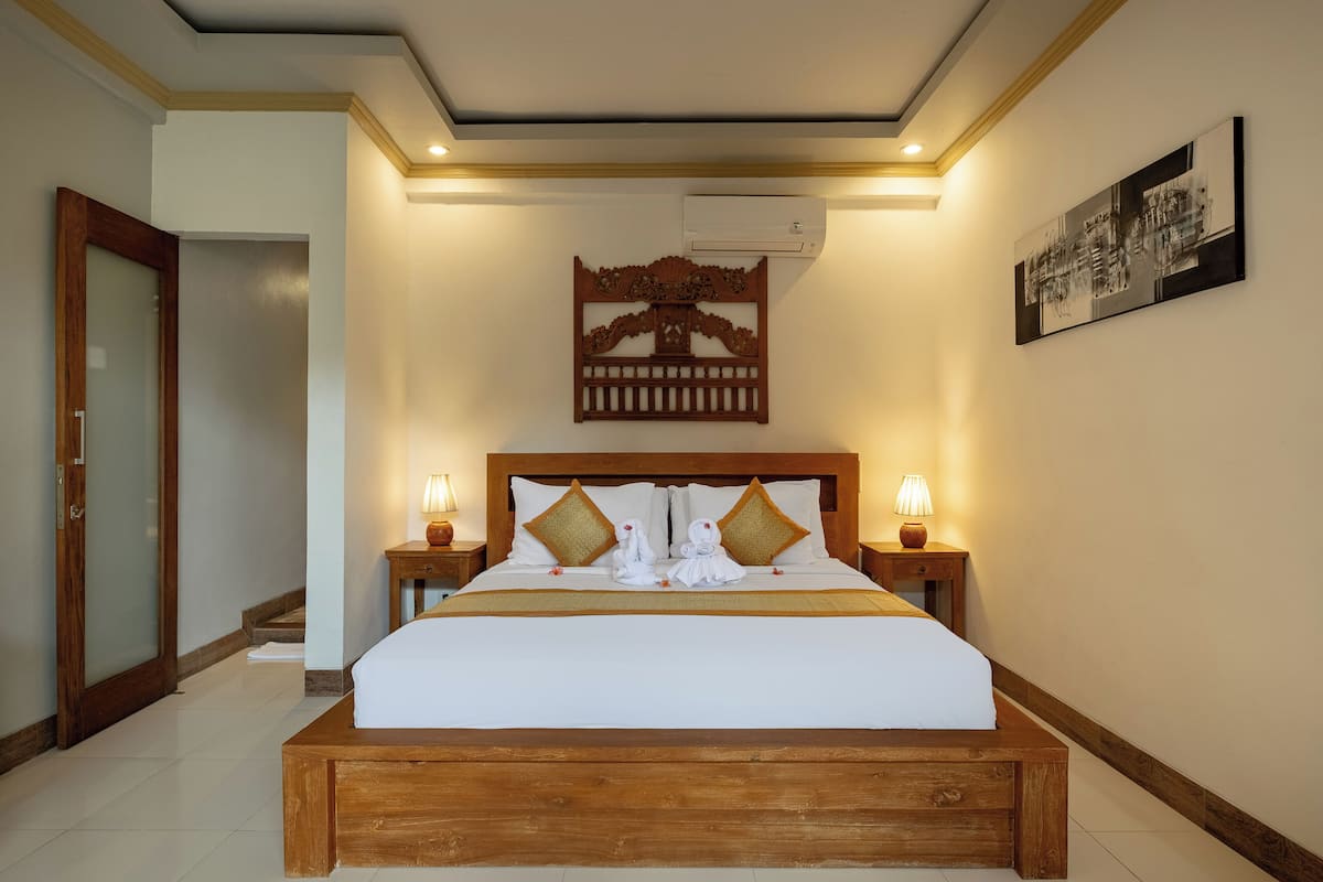 Habitación de hotel con una cama grande, cabecera de madera, dos mesitas de noche con lámparas, aire acondicionado de pared y una obra de arte enmarcada.