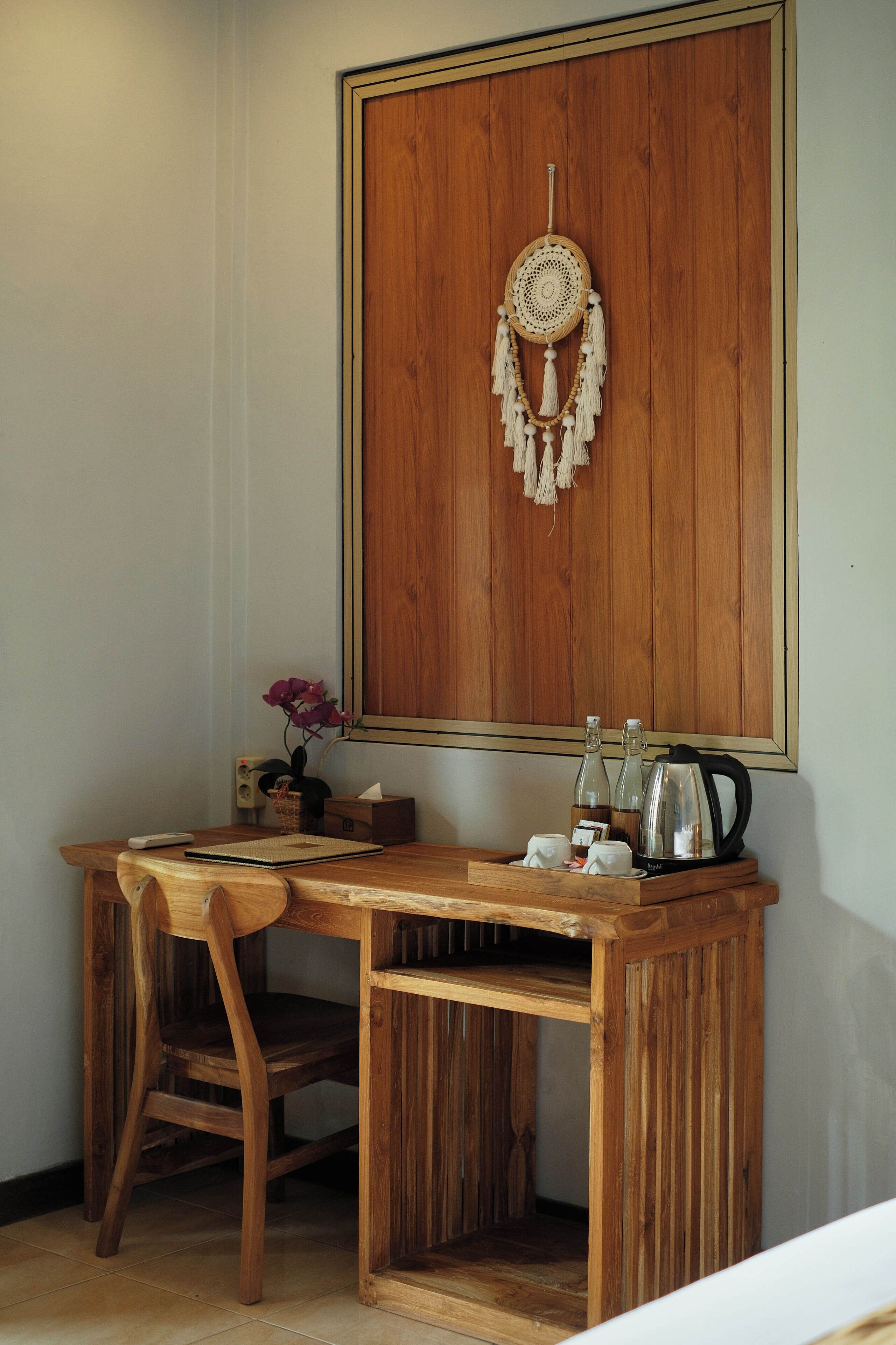 A wooden desk with a chair, a dreamcatcher, a vase with flowers, and a kettle.