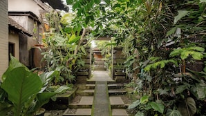 A narrow stone pathway flanked by lush greenery and potted plants, leading to a traditional building with a tiled roof. - Togog Polos Ubud House (Ubud)
