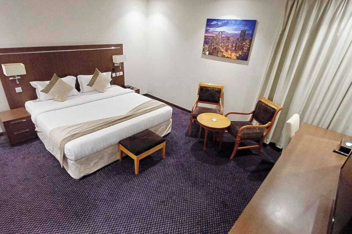 A hotel room with a large bed, a small round table, two chairs, a nightstand, and a painting on the wall.