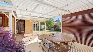 Un patio al aire libre con una mesa y sillas de madera, un área cubierta con luces de cuerda y un muro de ladrillo.