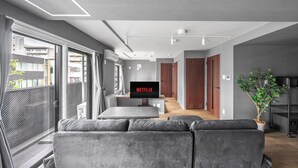 Een moderne woonkamer met een grijze bank, een flatscreen televisie waarop 'NETFLIX' staat, en een groot raam met uitzicht op gebouwen.