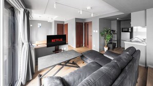 Een moderne woonkamer met een grijze bank, een televisie waarop 'NETFLIX' wordt weergegeven, en een eethoek met een tafel en stoelen.