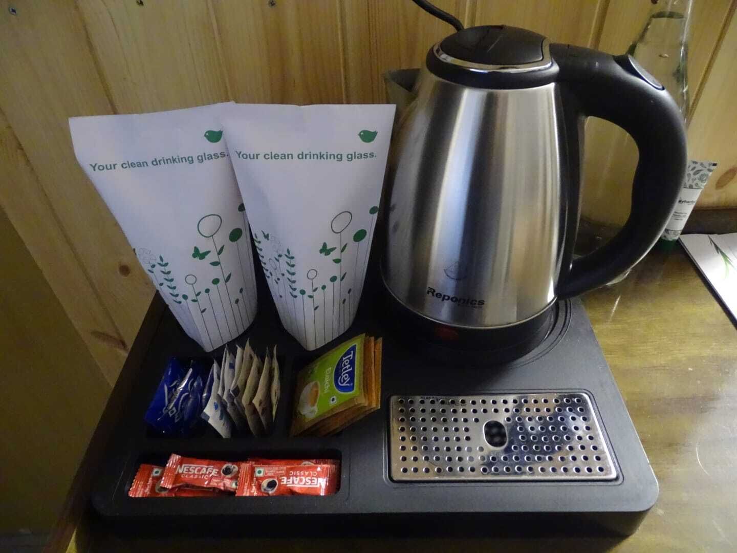 A stainless steel coffee pot, two paper cups with a green design, a pack of tissues, and a box of Nescafe Classic coffee on a black tray.