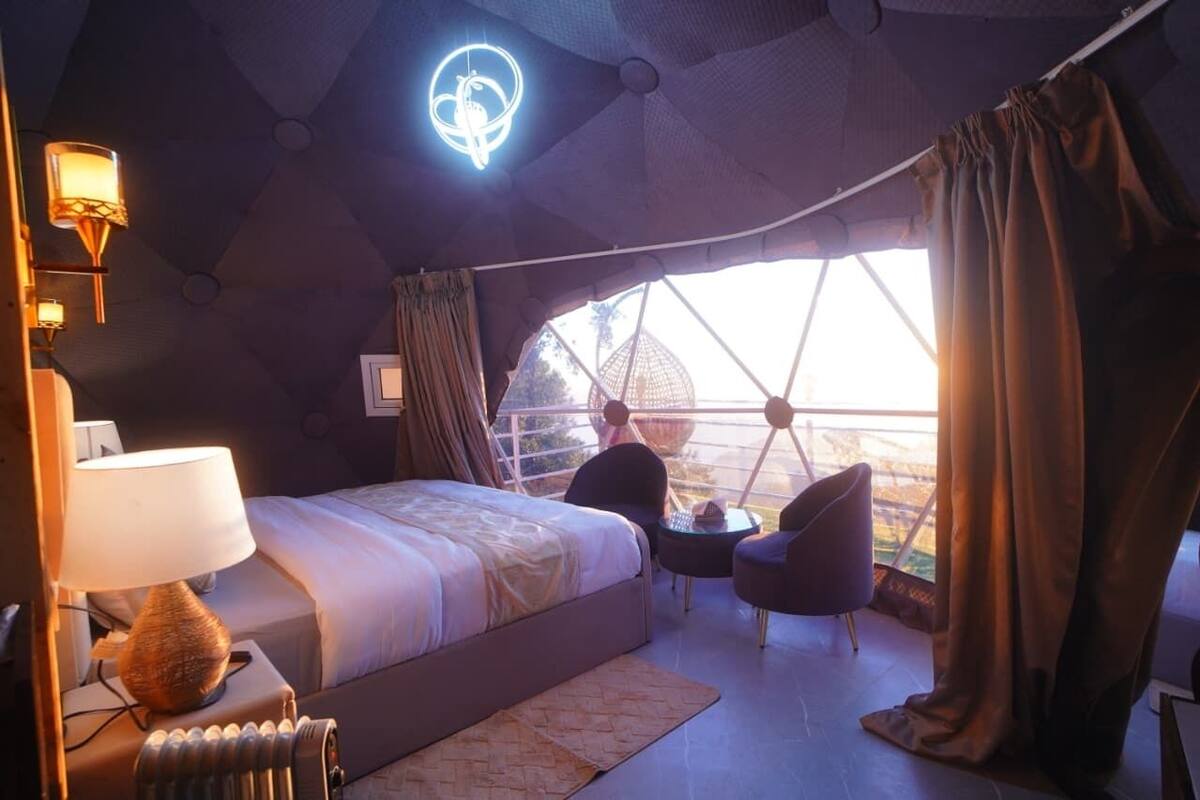 A modern bedroom with a large bed, a small table, two chairs, and a view of the outdoors through a large window.