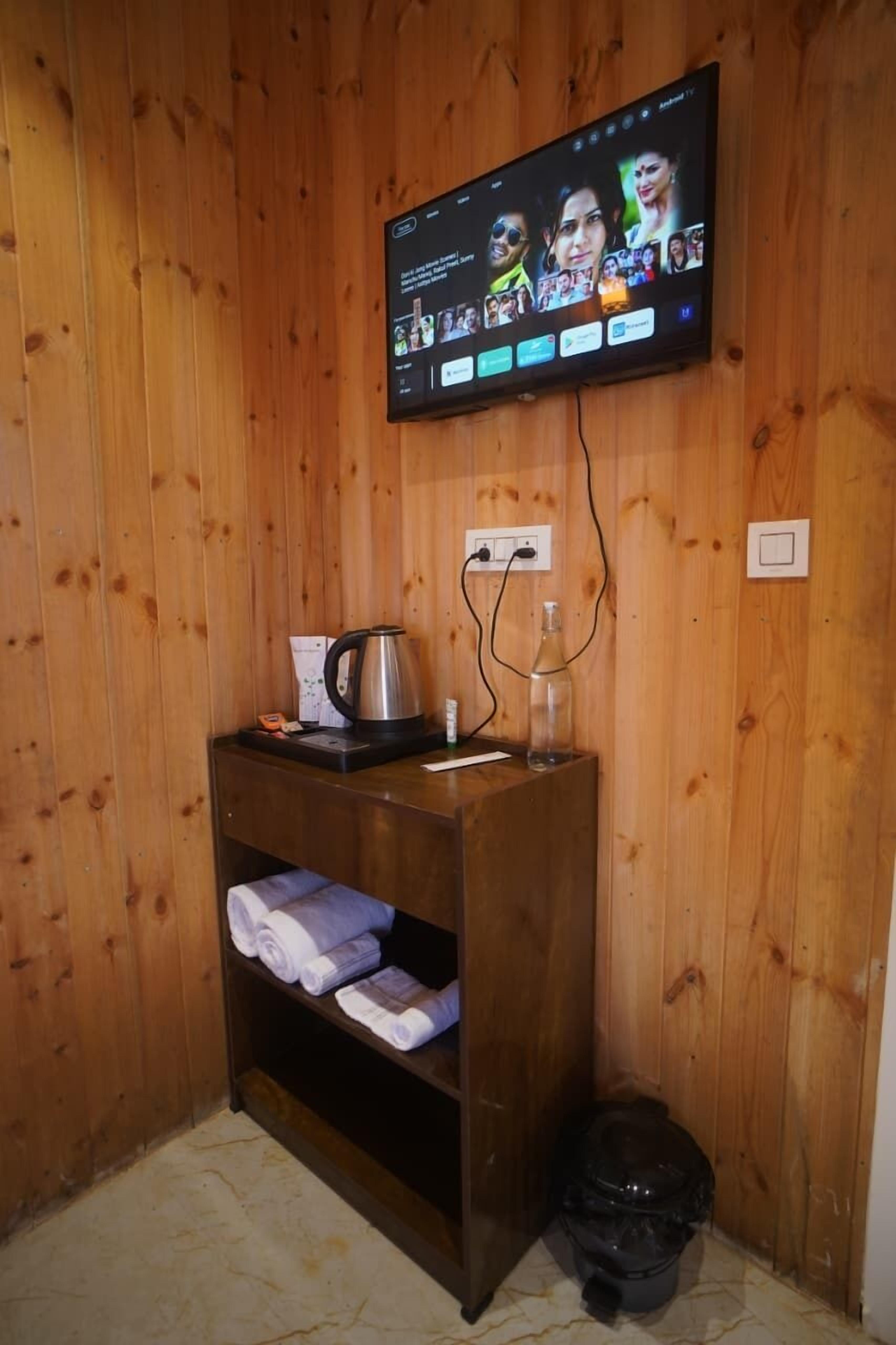 A wooden-paneled room with a TV mounted on the wall displaying a social media app. There is a small wooden table with a kettle, a bottle, and a cup. A trash bin is visible in the corner.