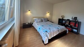 Ein Schlafzimmer mit einem Bett, Nachttischen, einem Wandregal mit Büchern und Dekoration sowie einem Fenster mit Vorhängen.