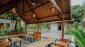An outdoor patio area with wooden tables and chairs, a stone pathway, and a covered seating area with hanging lights. - Nuansa Penida Cottage (Penida Island)