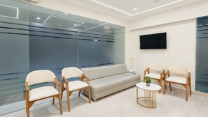 A modern waiting area with a sofa, wooden chairs, a small table, and a TV mounted on the wall. - Treebo Premium Bizz (Ahmedabad)
