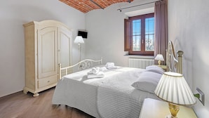 Un dormitorio con techo de ladrillo, un armario de madera, una cama con ropa blanca, una mesita de noche con lámpara y una ventana con cortinas.