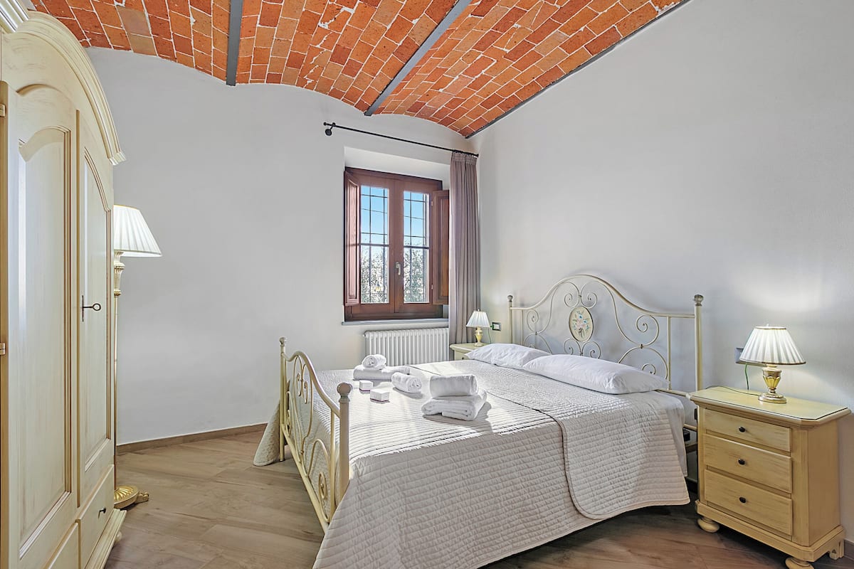 A bedroom with a brick ceiling, a large window with curtains, a bed with a white quilt, a bedside table with a lamp, and a wardrobe.