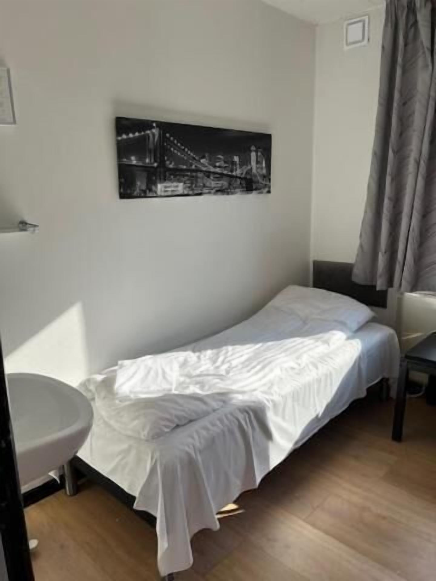 A single bed with a white bedspread in a room with a wooden floor, a small table, and a wall-mounted picture of a bridge.