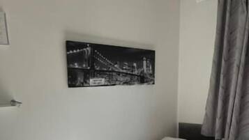 Uma cama de solteiro com edredom branco em quarto com piso de madeira, uma pequena mesa e um quadro pendurado na parede retratando uma ponte.