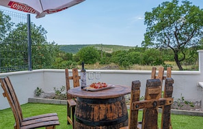 A rustic outdoor seating area with a wooden barrel table, chairs, and a wine bottle.