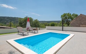 A rectangular swimming pool with a white border, surrounded by light-colored tiles. There are three lounge chairs and a red and white umbrella. The background features a hilly landscape with trees and a clear sky.