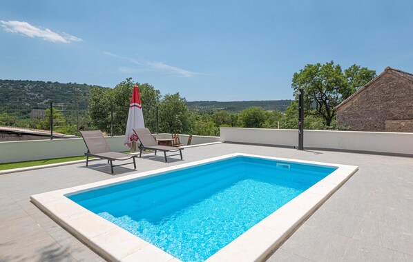 A rectangular swimming pool with a white border, surrounded by light-colored tiles. There are three lounge chairs and a red and white umbrella. The background features a hilly landscape with trees and a clear sky.