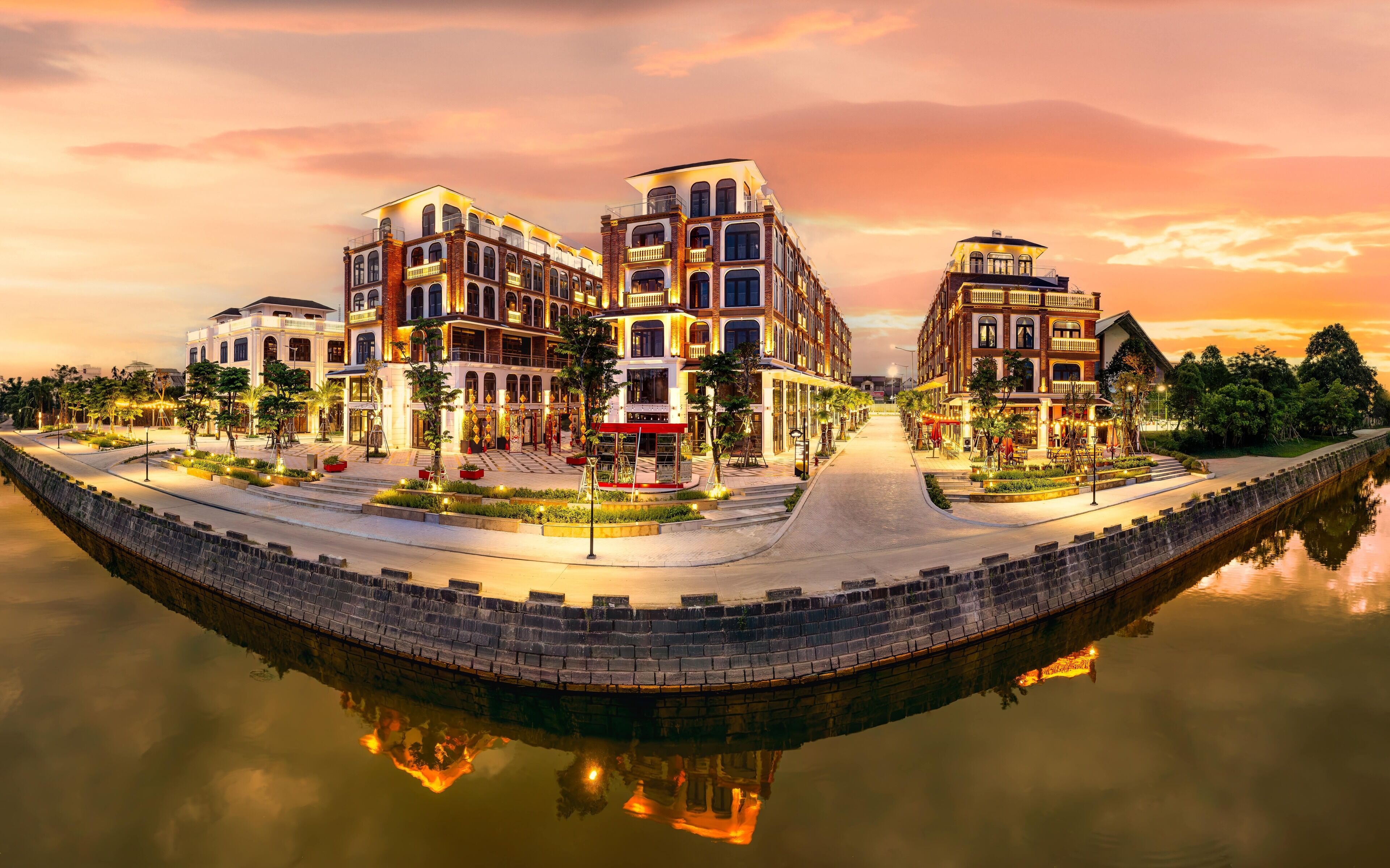 A waterfront hotel with multiple buildings, a reflective water body, and a well-lit outdoor area with trees and seating.