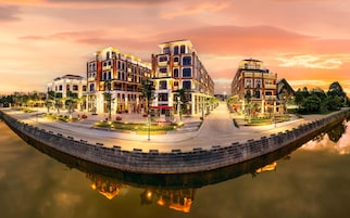 Un hotel frente al agua con varias edificaciones, una superficie acuática reflectante y un área exterior iluminada con árboles y asientos.