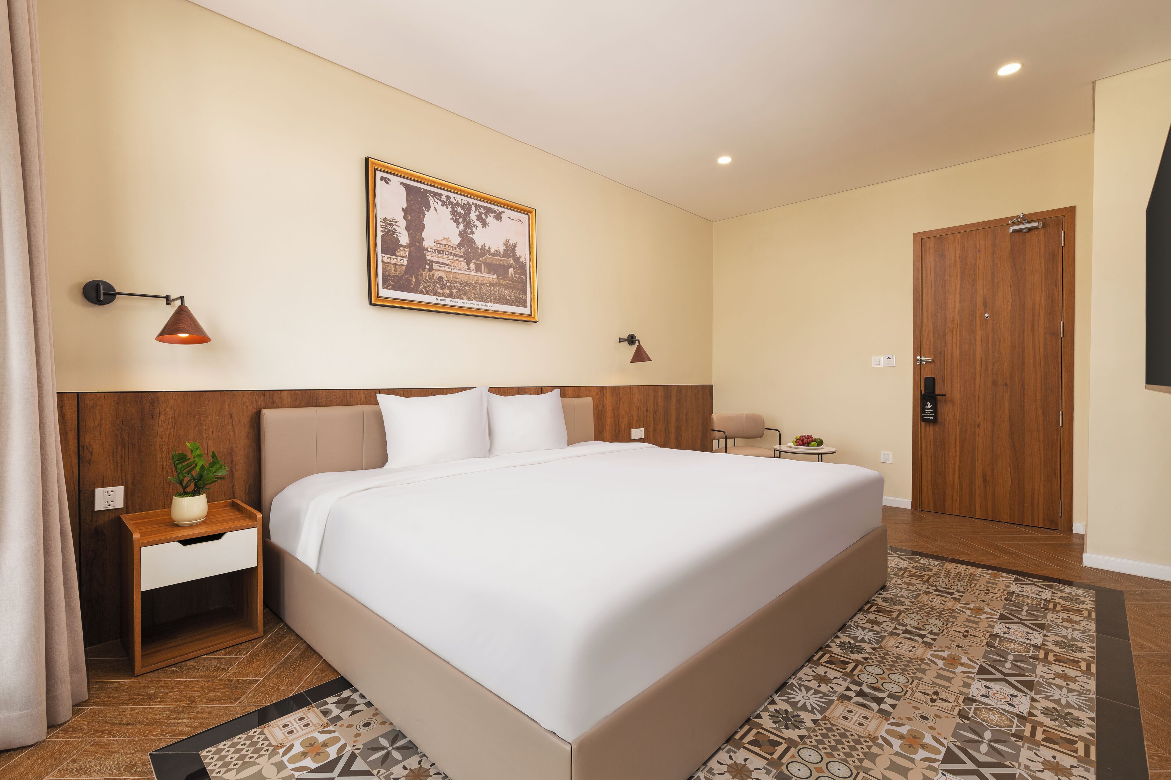 A hotel room with a large bed, a nightstand with a plant, a framed picture on the wall, and a wooden door.