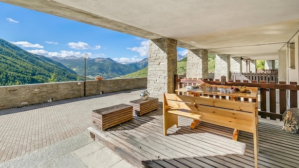 Un espacio al aire libre con techo, con pilares de piedra, bancos de madera y vistas a las montañas.