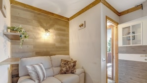 Una sala de estar acogedora con un sofá, un tapete blanco y esponjoso, una pared de madera, una planta colgante y un aplique de pared.