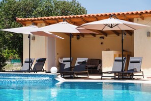 A swimming pool with lounge chairs and umbrellas, a tiled deck, and a building with a tiled roof. - Villa Ioannis (Mandra Eidyllia)