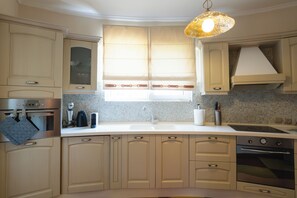 A kitchen with light-colored cabinets, a double sink, a stove, and a hanging light fixture. - Villa Ioannis (Mandra Eidyllia)