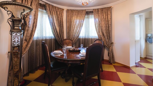 Een eetkamer met een ronde tafel voor vier personen, omringd door stoelen, een kroonluchter en gordijnen.