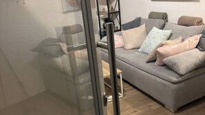 A modern living room with a grey sofa, multiple cushions, and a glass door leading to another room. - Emsquartier 2 (Emsdetten)