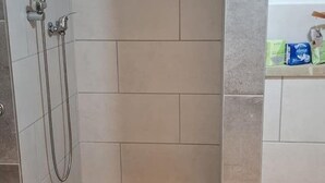 A bathroom with beige tiles, a shower with a handheld hose, and a shelf with cleaning products. - Emsquartier 1 (Emsdetten)