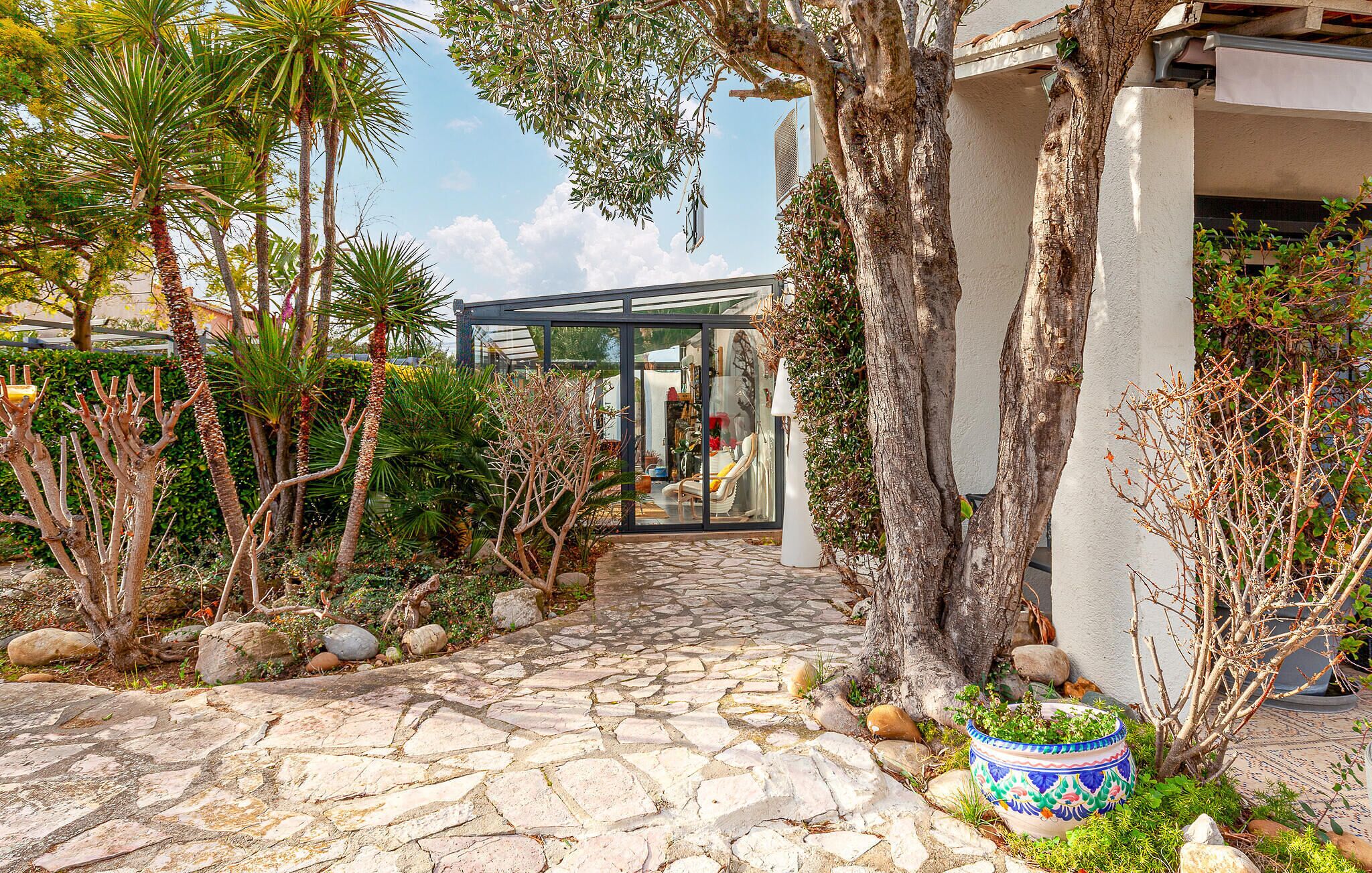 A stone-paved pathway leading to a modern glass structure, surrounded by various plants and trees.