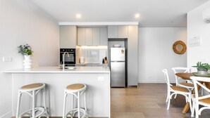 A modern kitchen with white cabinetry, a built-in oven, and a stainless steel refrigerator. There are two bar stools with woven seats and a dining table with white chairs. - Preston Rooftop Oasis - Pool, Views & City Access (Preston)