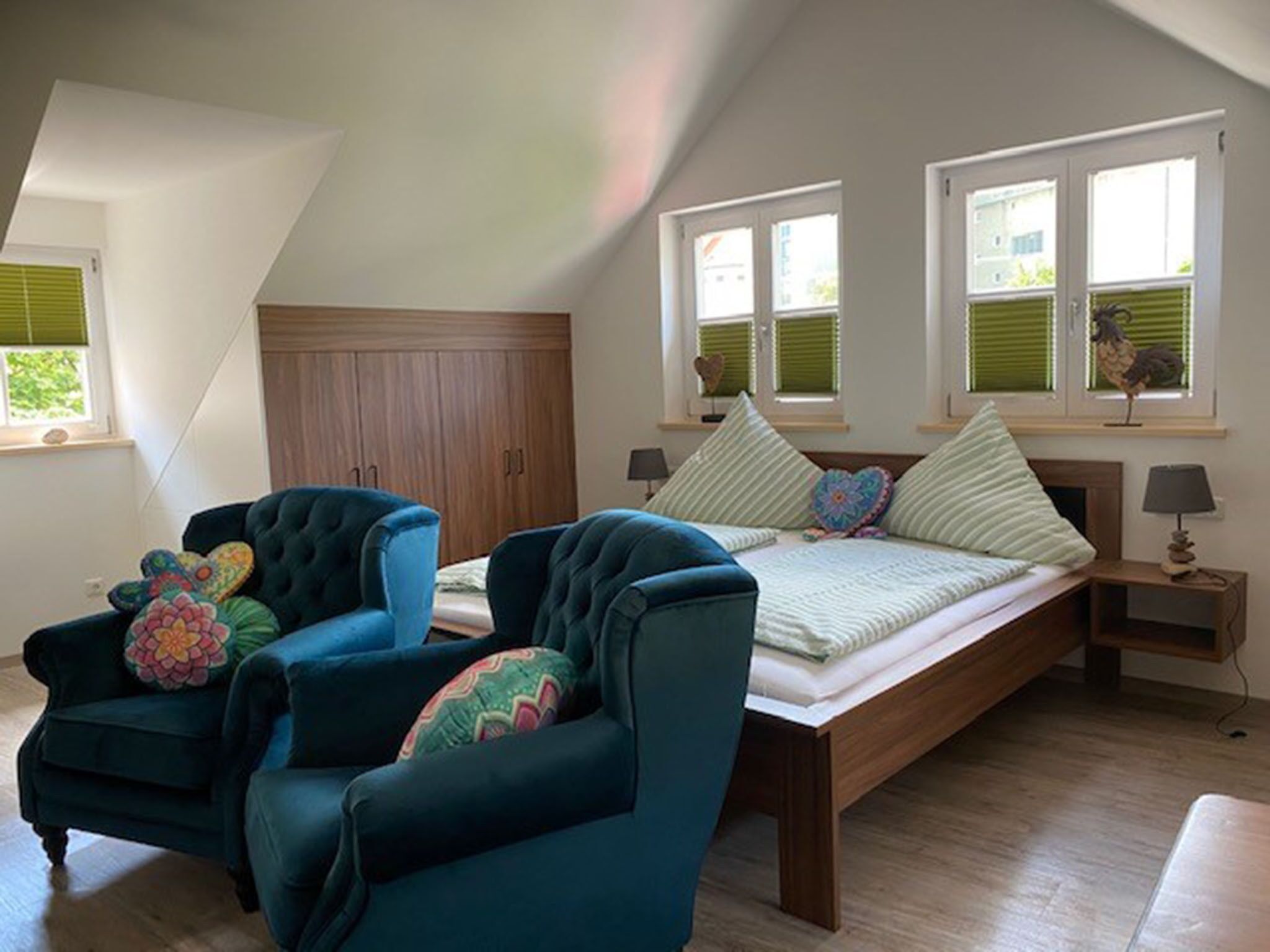 A bedroom with a loft bed, two armchairs, a wooden wardrobe, and a window with blinds.