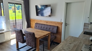 Een zithoek met een houten tafel en beklede stoelen, een televisie boven de tafel en uitzicht op een groen landschap door het raam.