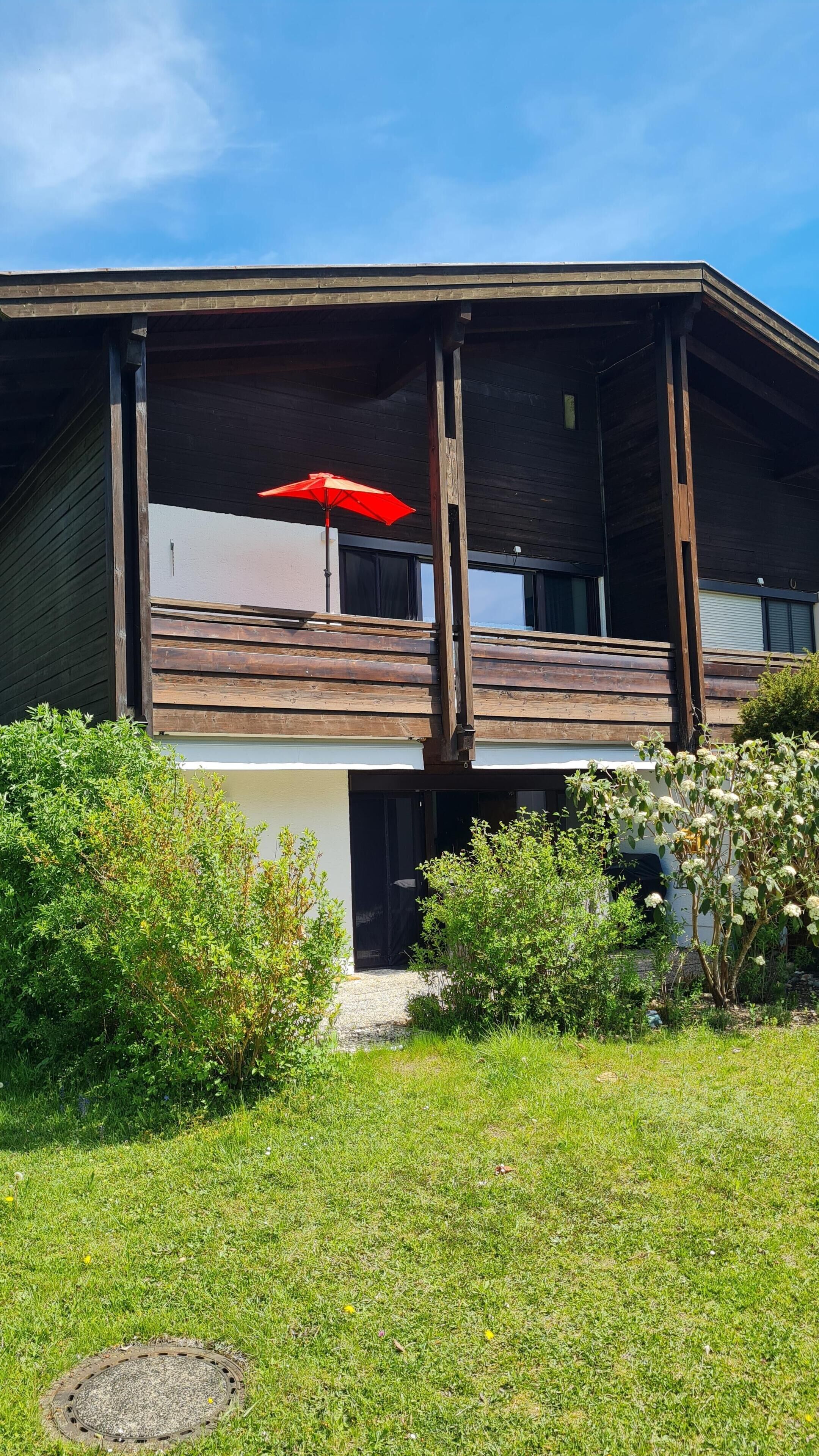 A two-story building with a wooden facade, a red umbrella, and a grassy yard.