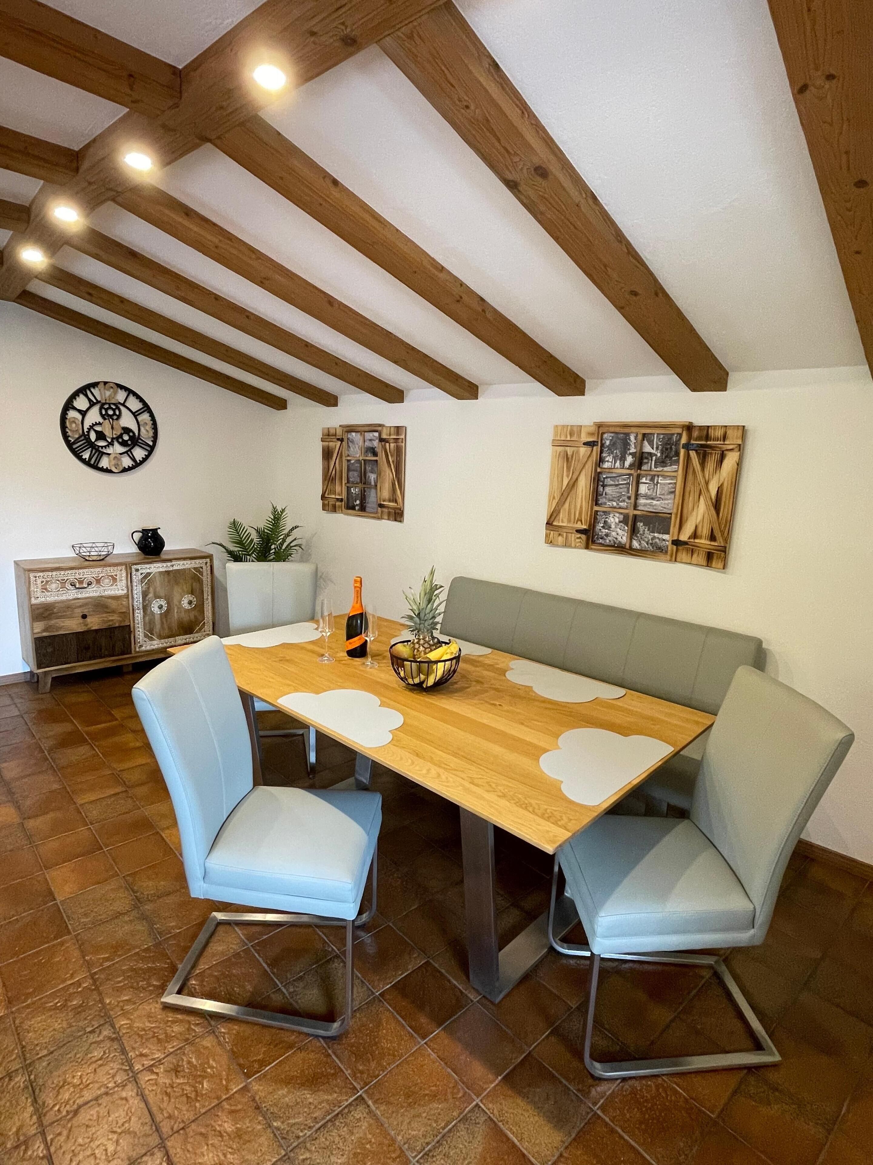 A dining area with a wooden table, two cushioned chairs, a bench, a decorative wall clock, and a cabinet with a plant.
