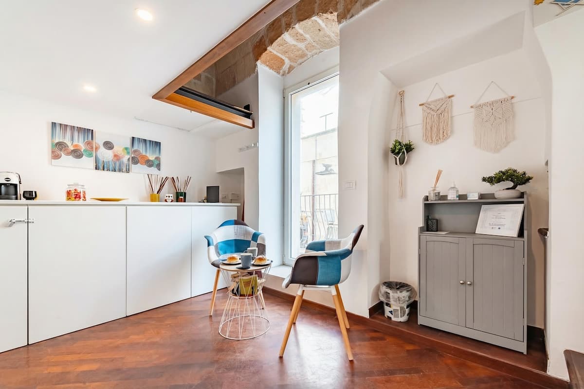 Una cocina moderna con armarios blancos, un rincón de comedor con dos sillas y un mueble gris con una planta.