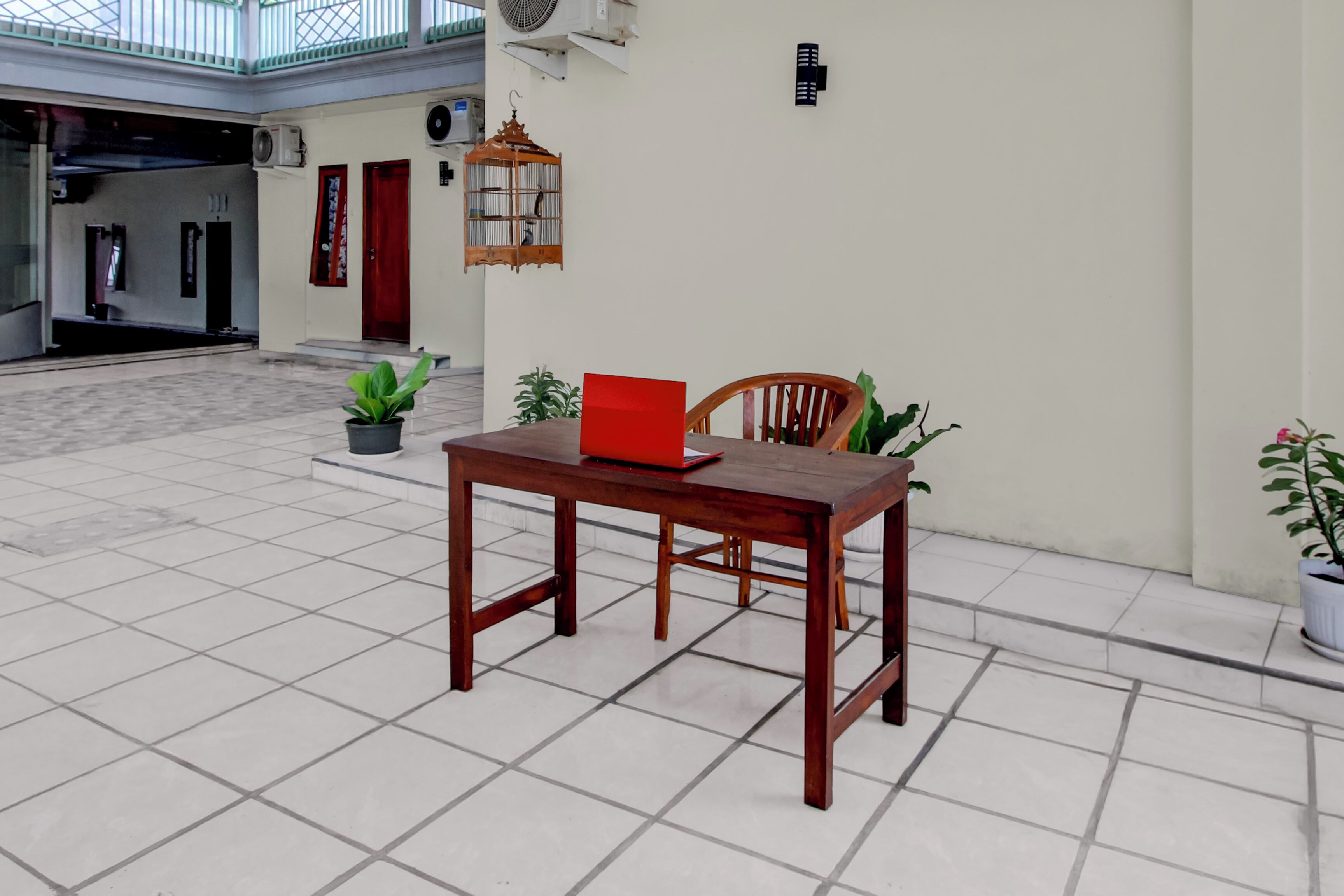 A wooden table with a red folder, a chair, and potted plants in a spacious indoor area with tiled flooring and a high ceiling.