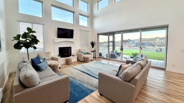 A spacious living room with a fireplace, large windows, and a view of the outdoors. - Luxury Home at Bear Lake! Sleeps 30! Resort Hot Tub! Tesla Charging! (Fish Haven)