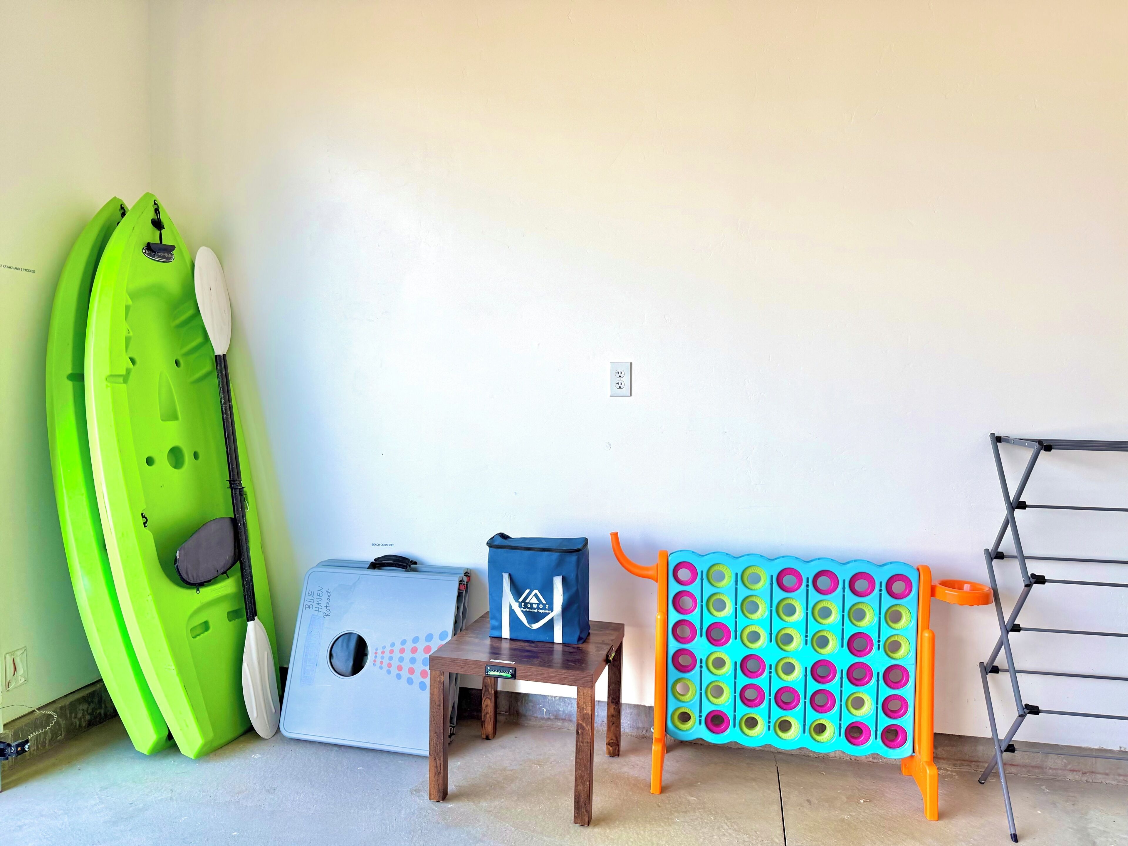 A room with a green kayak, a blue cooler, a small wooden table, and a colorful drying rack.
