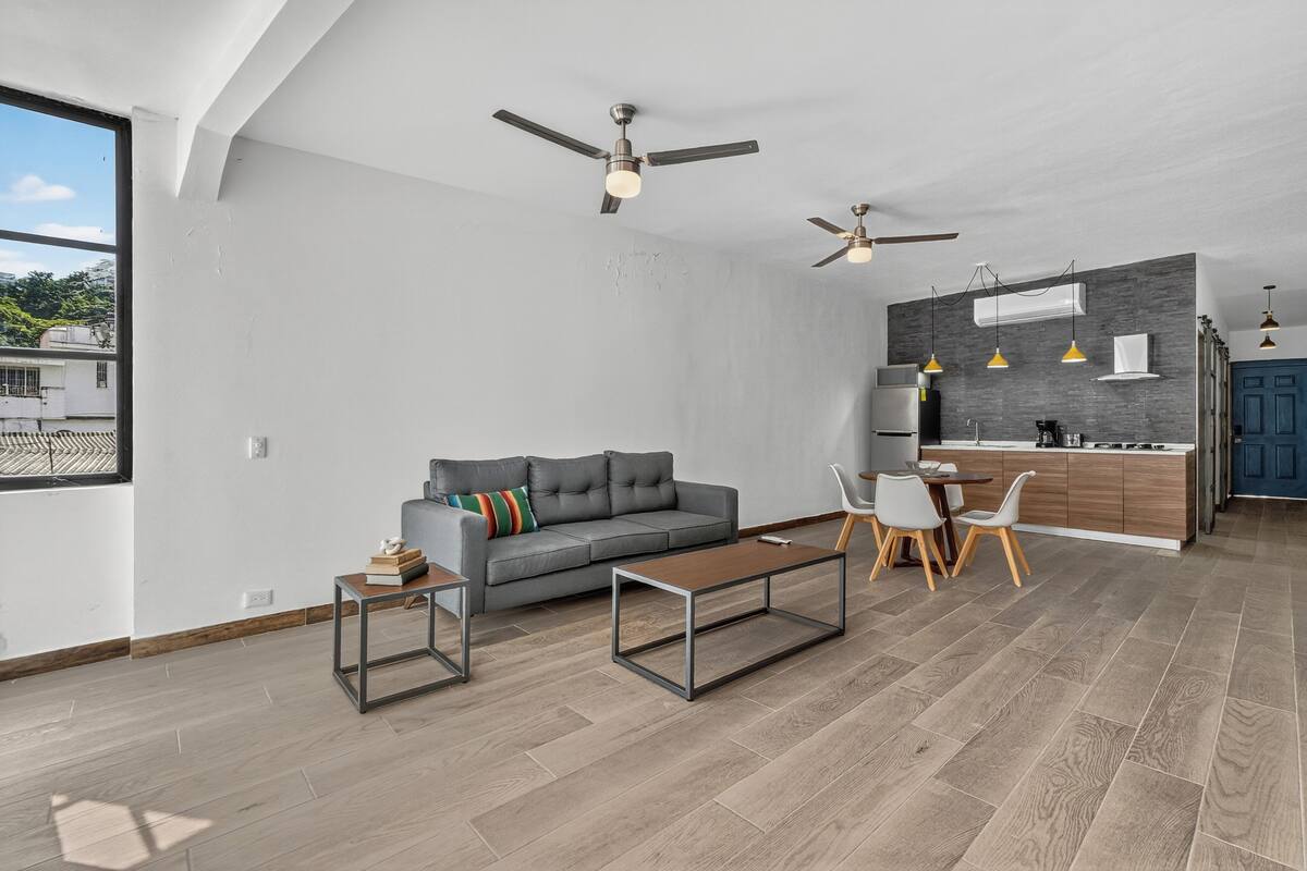 A modern living room with a gray sofa, a coffee table, and a dining area with chairs. There are ceiling fans and pendant lights.